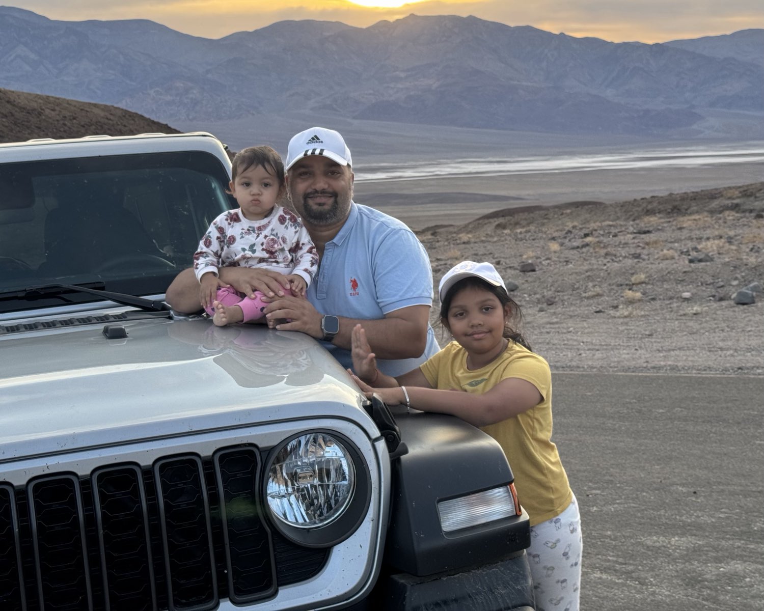 Abhishek Pathak with his two young daughters, leaning against the front of a jeep at sunset, with desert mountains and a dry lake bed behind them.